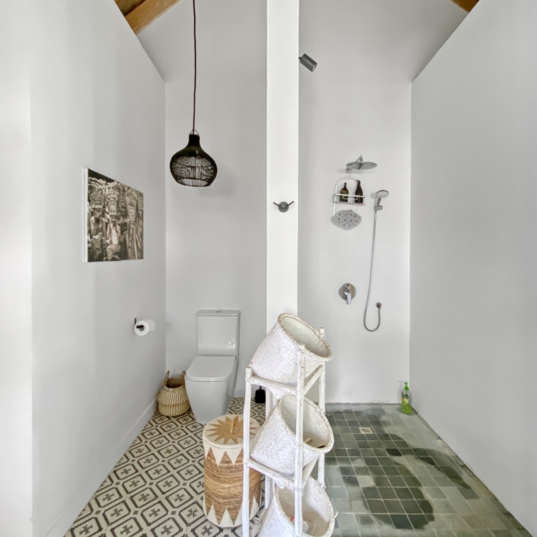 Bright narrow bathroom with white walls, a patterned floor, and a toilet on the left; a hanging black lantern-style light, wicker baskets, and a chair with folded towels along the right wall.