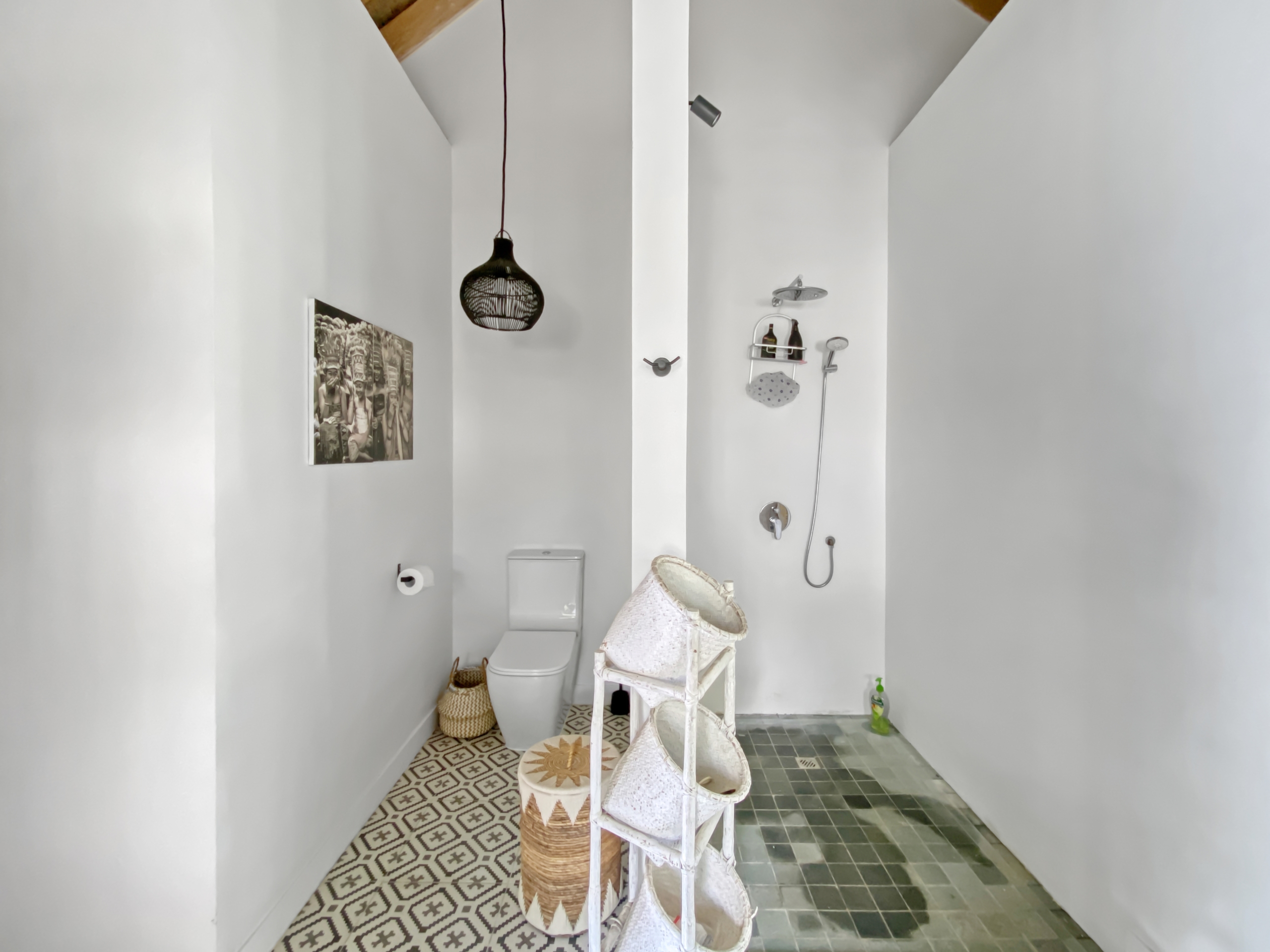 f19605cdc1c3384a772654962e8adee275fcd371 Bright narrow bathroom with white walls, a patterned floor, and a toilet on the left; a hanging black lantern-style light, wicker baskets, and a chair with folded towels along the right wall.