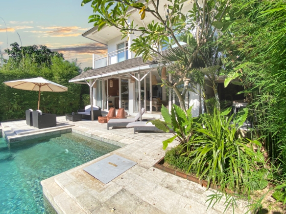 Sunny backyard with a rectangular pool, beige stone deck, wicker lounge chairs, and an umbrella beside a white house surrounded by tropical plants.