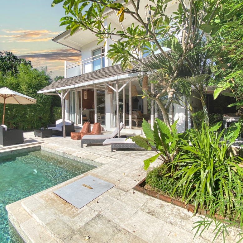 Sunny backyard with a rectangular pool, beige stone deck, wicker lounge chairs, and an umbrella beside a white house surrounded by tropical plants.