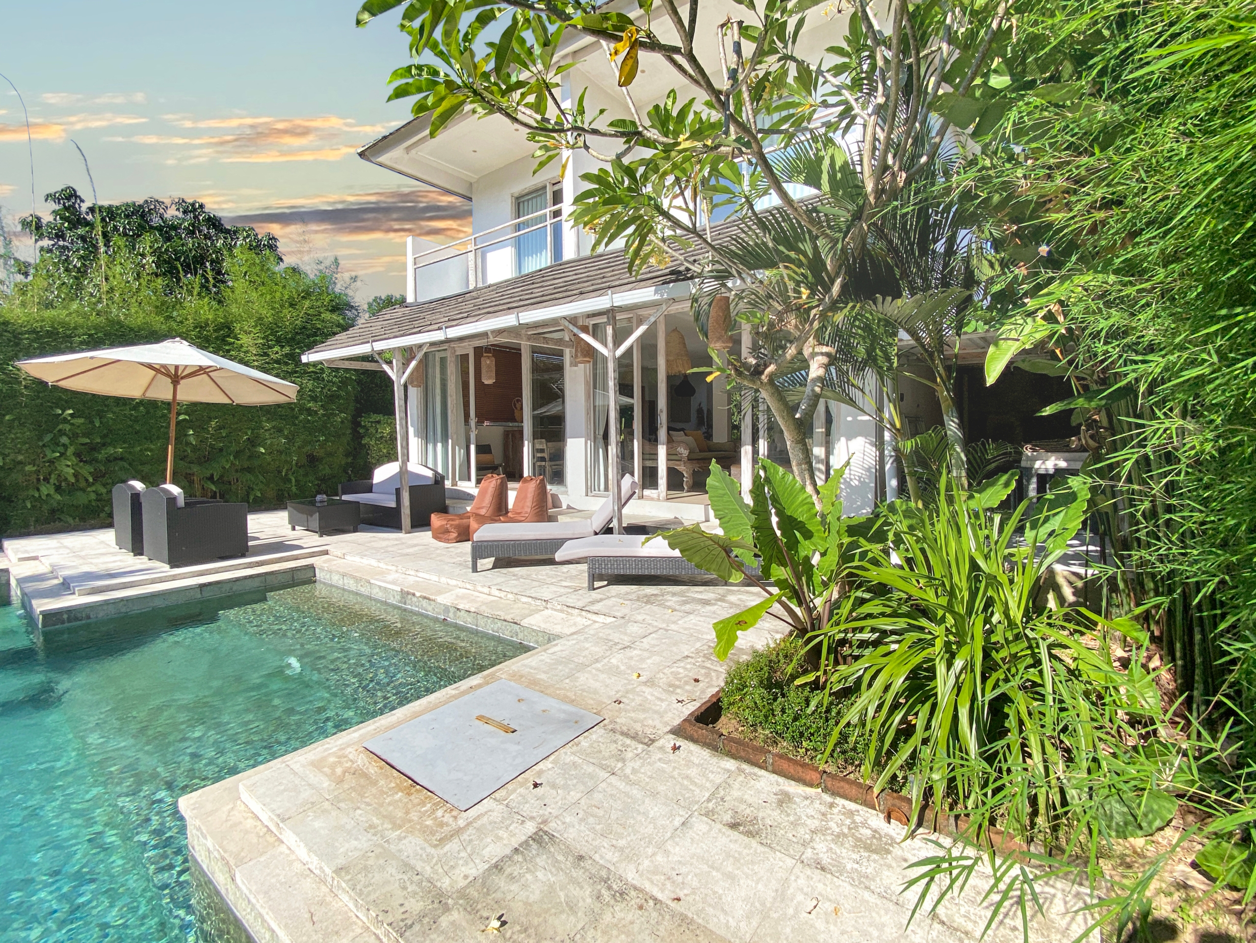 Sunny backyard with a rectangular pool, beige stone deck, wicker lounge chairs, and an umbrella beside a white house surrounded by tropical plants.