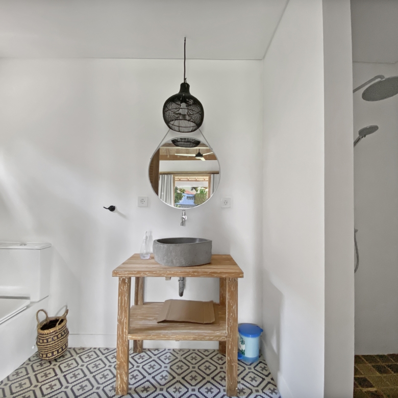 Bathroom with white walls, a wooden table supporting a gray stone sink, round mirror on the wall, and a black woven pendant light above; patterned tile floor.
