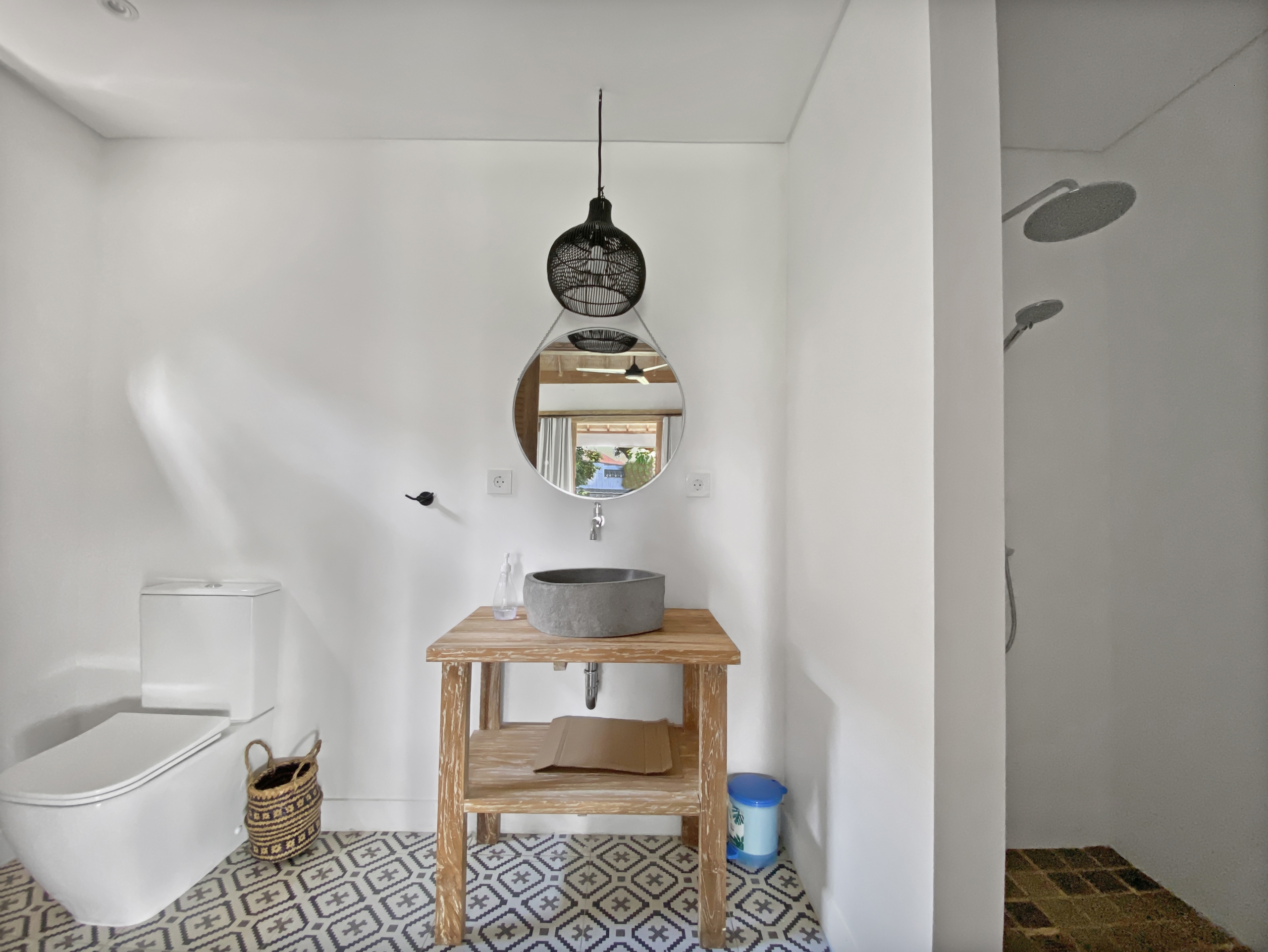 f4c0664d378bff67815d0b75d1e90bc5778a39b2 Bathroom with white walls, a wooden table supporting a gray stone sink, round mirror on the wall, and a black woven pendant light above; patterned tile floor.