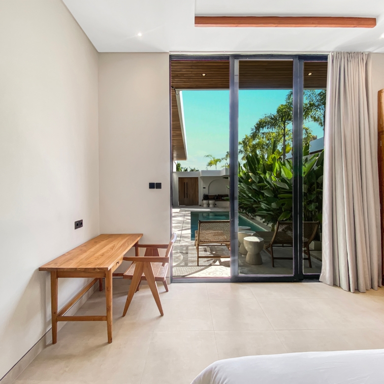 Hotel room interior with a wooden desk and bench along a beige wall, sliding glass doors to a patio with tropical plants outside.