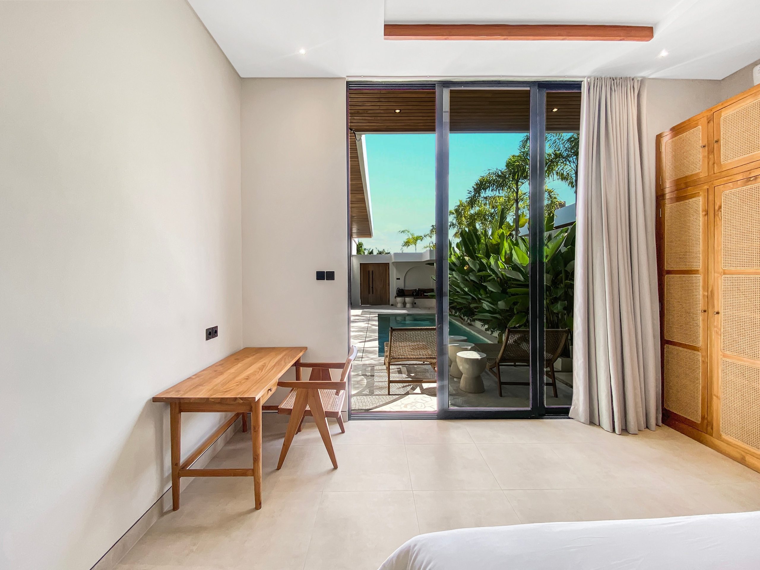 Hotel room interior with a wooden desk and bench along a beige wall, sliding glass doors to a patio with tropical plants outside.