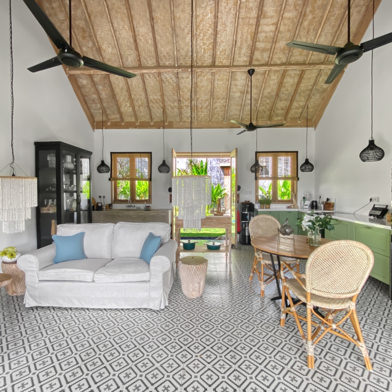 Open-plan living area with a gray sofa and blue cushions, woven chairs, and a patterned tile floor under a wood-beamed ceiling with ceiling fans.