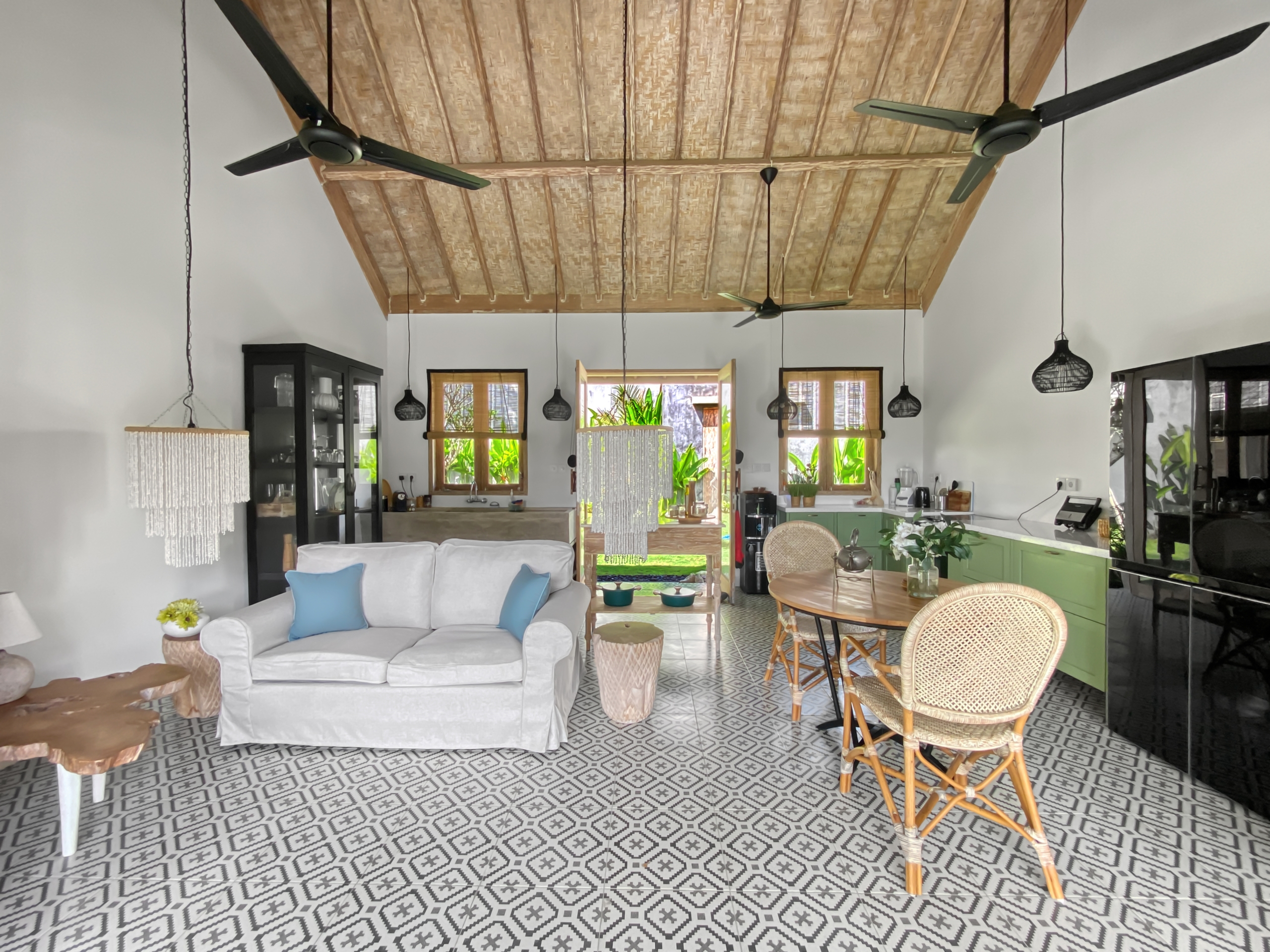 ffdb102ded2e65ad9d17dab0f68ba43675e4764f Open-plan living area with a gray sofa and blue cushions, woven chairs, and a patterned tile floor under a wood-beamed ceiling with ceiling fans.