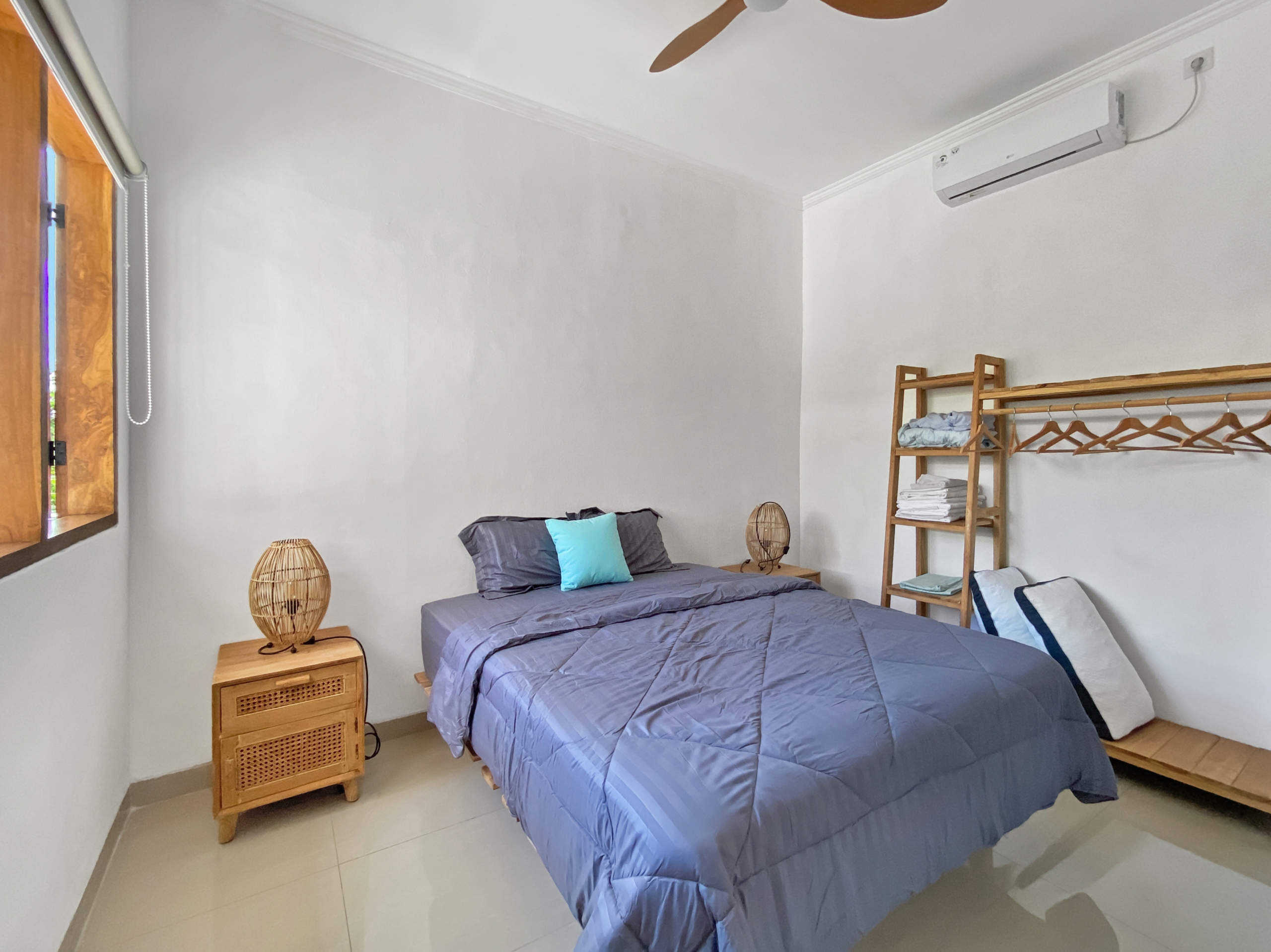 0106ce97c337e203e6ae2beeb12e4bb61af1e81d Twin bedroom with two single beds in blue comforters, a wooden nightstand with a wicker lamp, and a wall-mounted clothes rack.