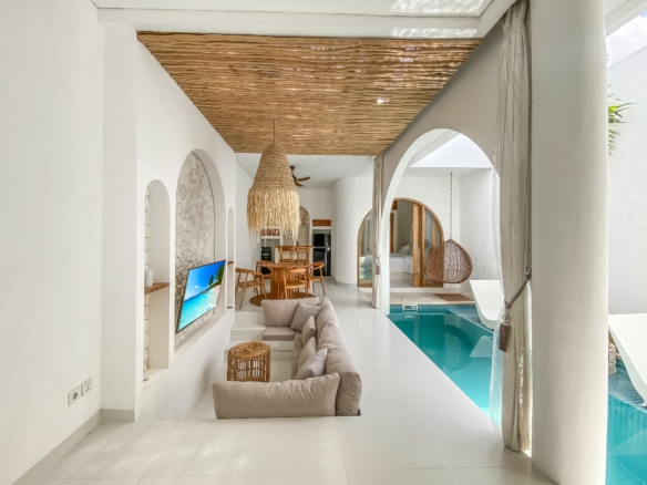 Open living area with white walls, arched doorways, a large woven pendant light, wooden dining set, and a view of the indoor pool.