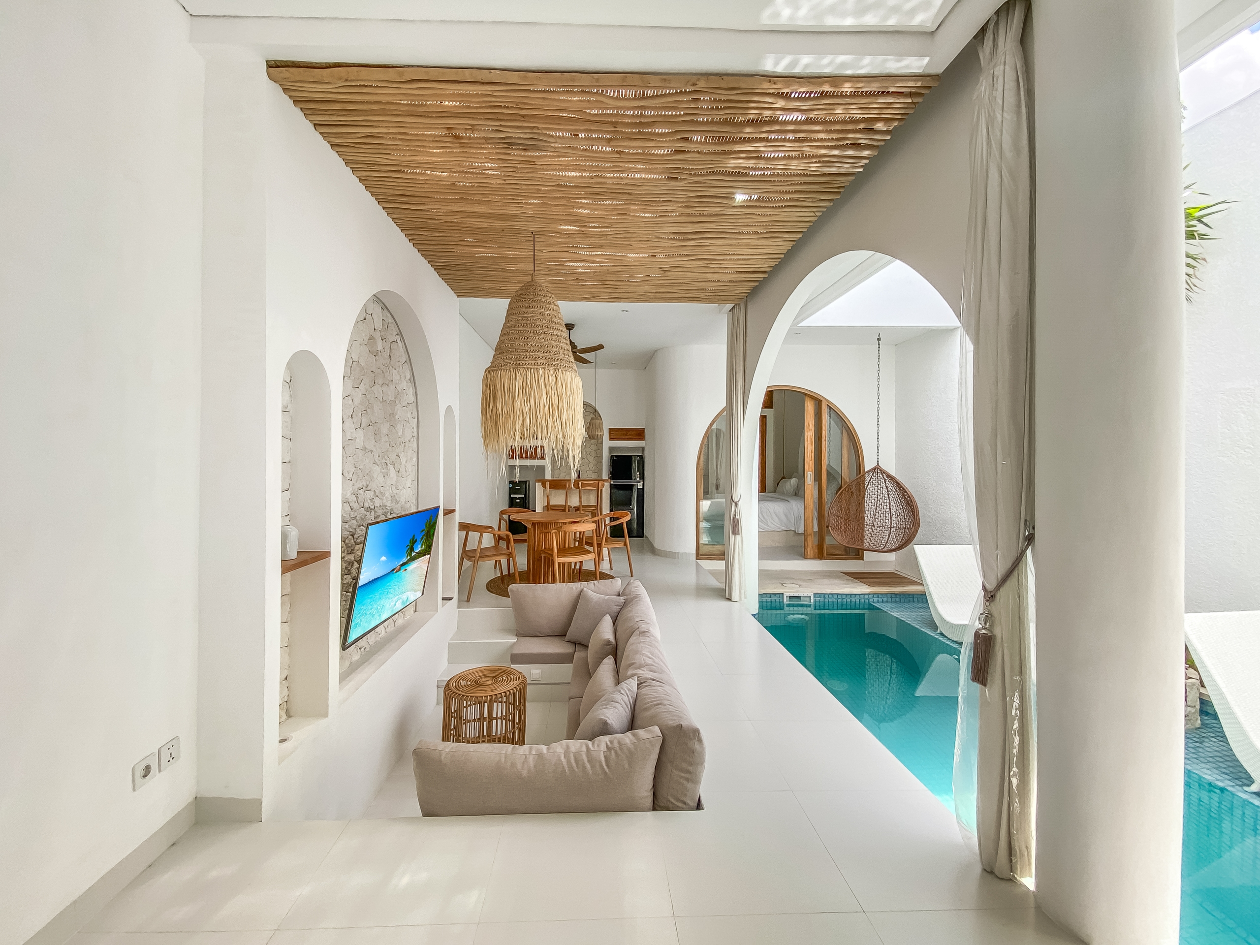 0370aea7874bce98be1b4d44248c75fbb132363e Open living area with white walls, arched doorways, a large woven pendant light, wooden dining set, and a view of the indoor pool.