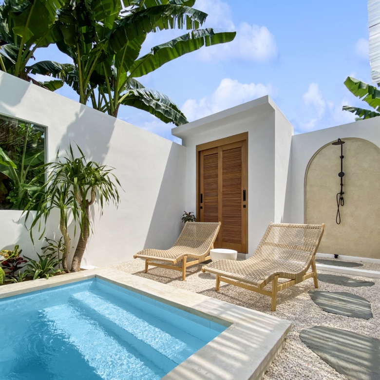 Poolside courtyard with white walls, beige wicker lounge chairs, and banana trees under a blue sky.