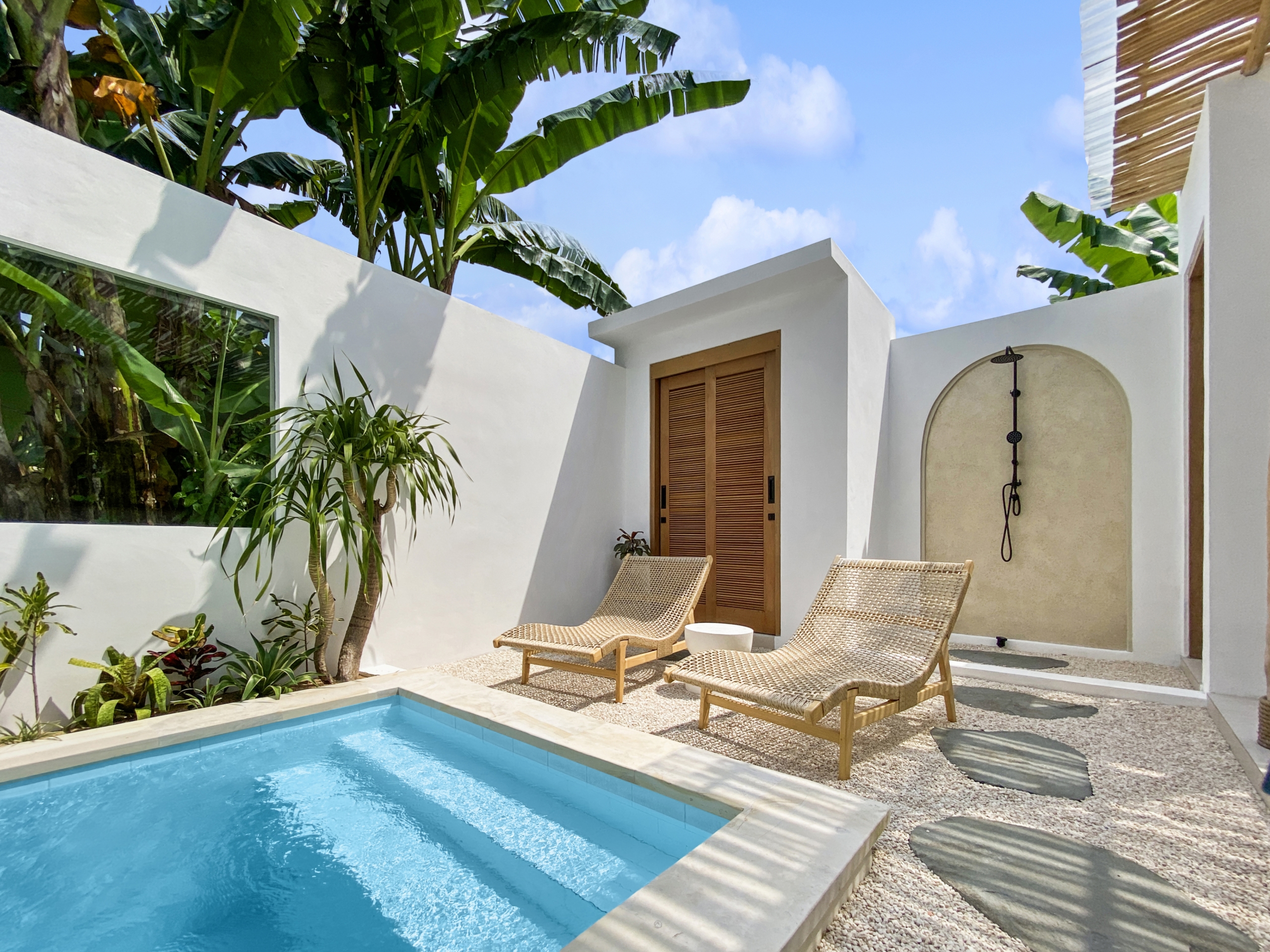 08c2ea69ff88668eb753936c6d1ac91a75892fa3_2306-68d5cfb10ffde Poolside courtyard with white walls, beige wicker lounge chairs, and banana trees under a blue sky.