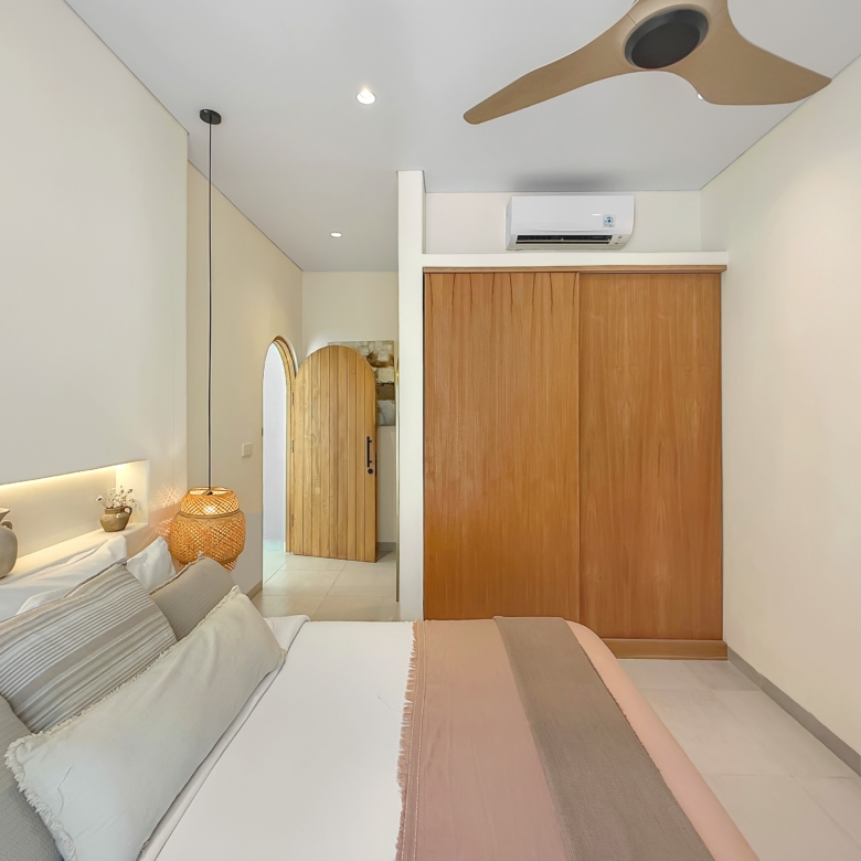 Two neatly made twin beds with beige bedding and decorative cushions in a modern bedroom, next to a wooden wardrobe and warm lighting.