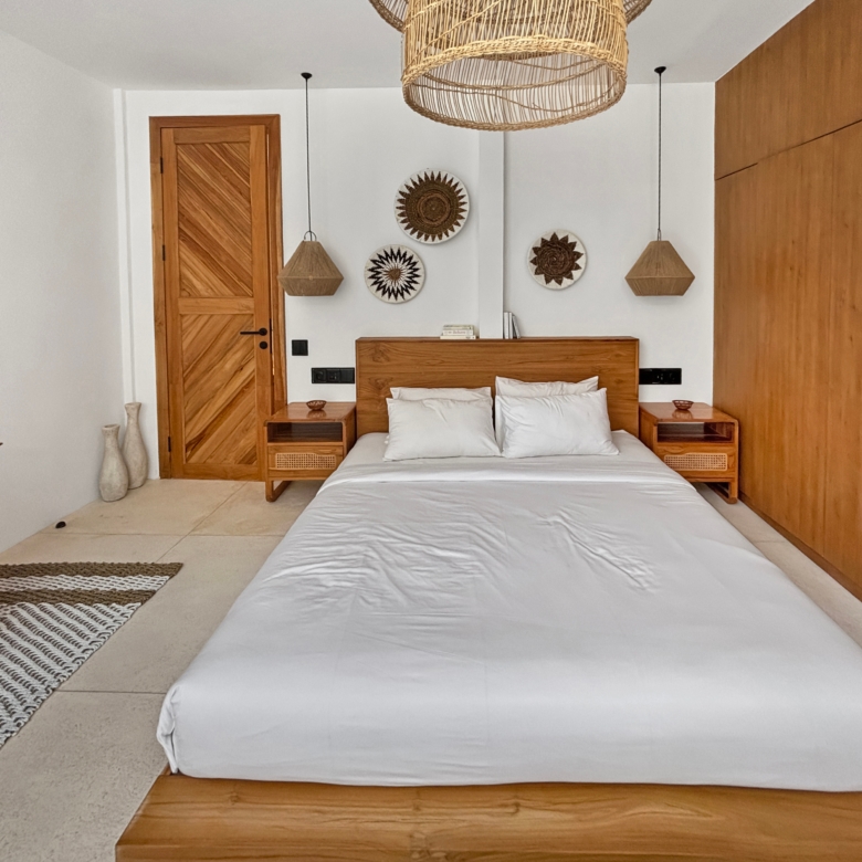 Wooden-framed bed with white bedding, two nightstands, and woven pendant lights in a bright white room with wooden accents.