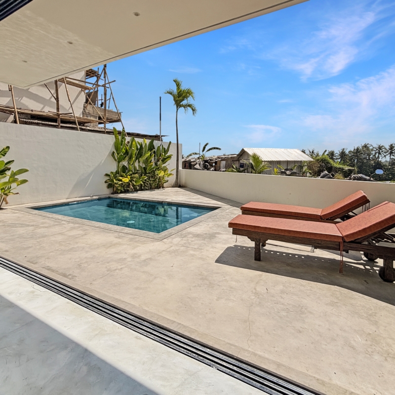 View from inside a glass-walled room onto a sunny poolside patio with lounge chairs and tropical plants.