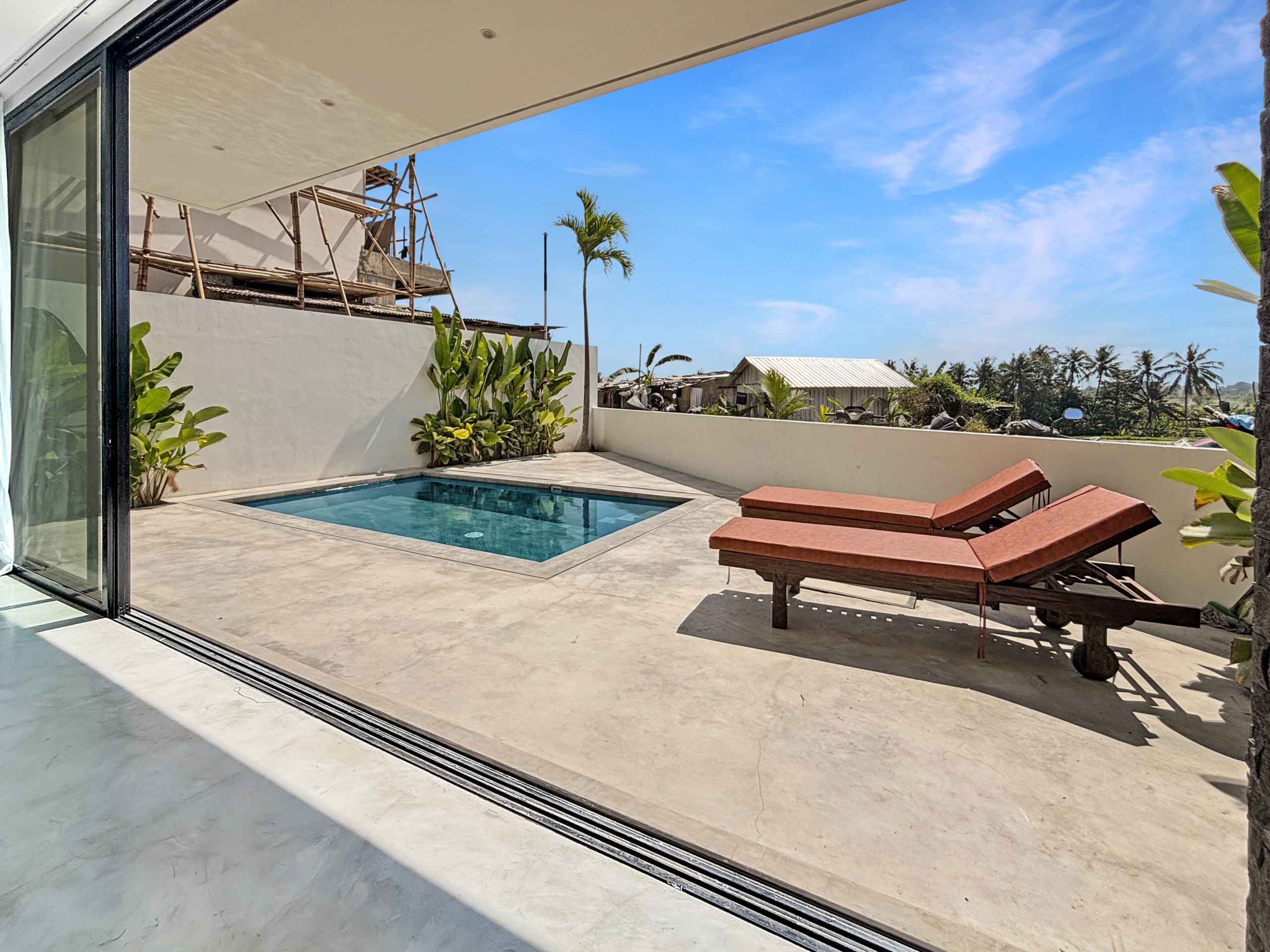 0c19b725326c051cb5e4bbb38f7fd978480d81aa_6795-6a1d7bad5ccea View from inside a glass-walled room onto a sunny poolside patio with lounge chairs and tropical plants.