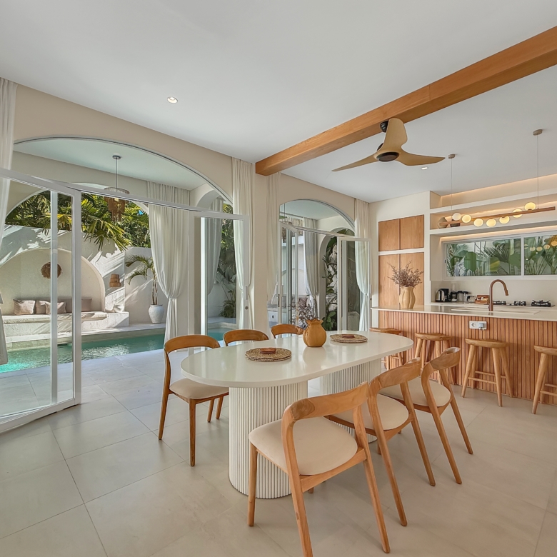 Open-plan kitchen and dining area with a white island, wooden stools, and glass doors opening to a pool and palm garden in the yard.