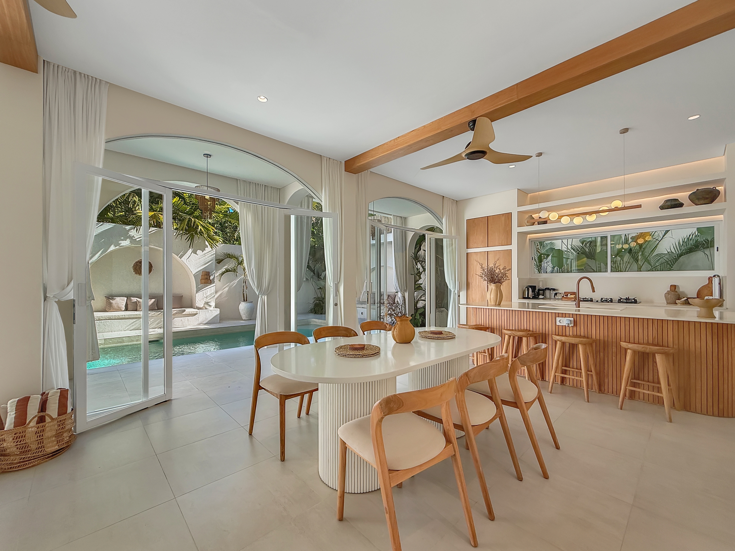 0e4c3e036270ceced54727893fad2b2b6004791c Open-plan kitchen and dining area with a white island, wooden stools, and glass doors opening to a pool and palm garden in the yard.