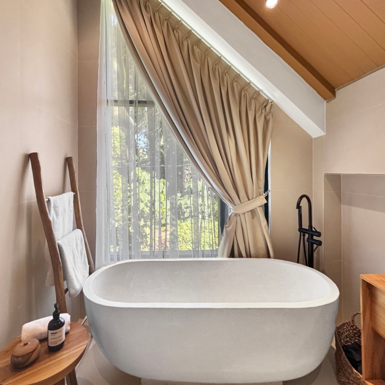 Freestanding white bathtub in a sunlit beige bathroom with sheer and draped curtains at the window. A wooden towel ladder and small table sit nearby.