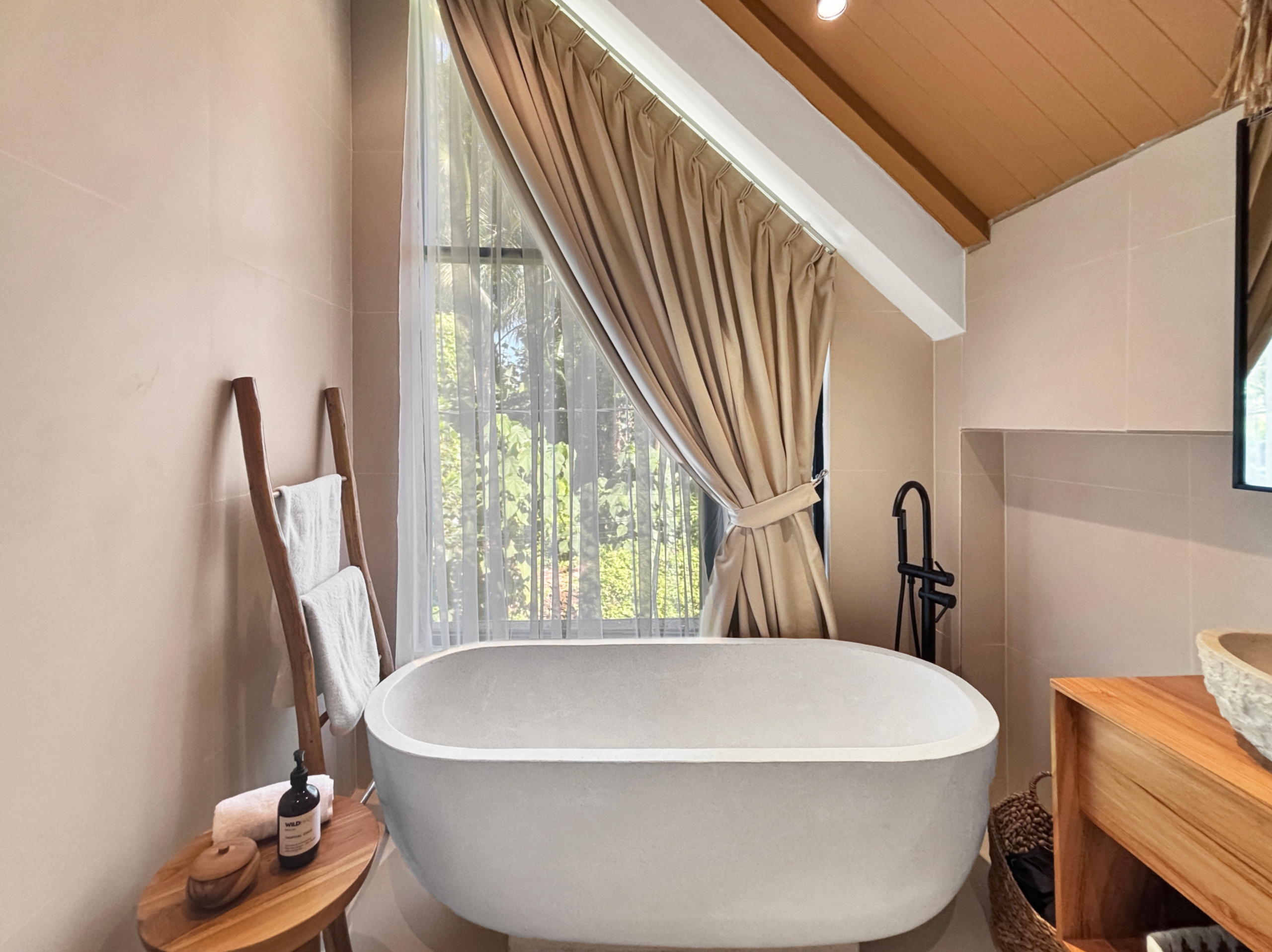 0ee04a8905747f61a07e982a034413df56070680 Freestanding white bathtub in a sunlit beige bathroom with sheer and draped curtains at the window. A wooden towel ladder and small table sit nearby.