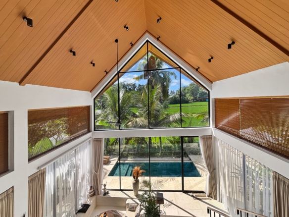 Interior view of a double-height living area featuring a wooden vaulted ceiling, black-framed triangular window, and palm trees outside