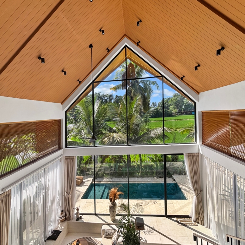 Interior view of a double-height living area featuring a wooden vaulted ceiling, black-framed triangular window, and palm trees outside