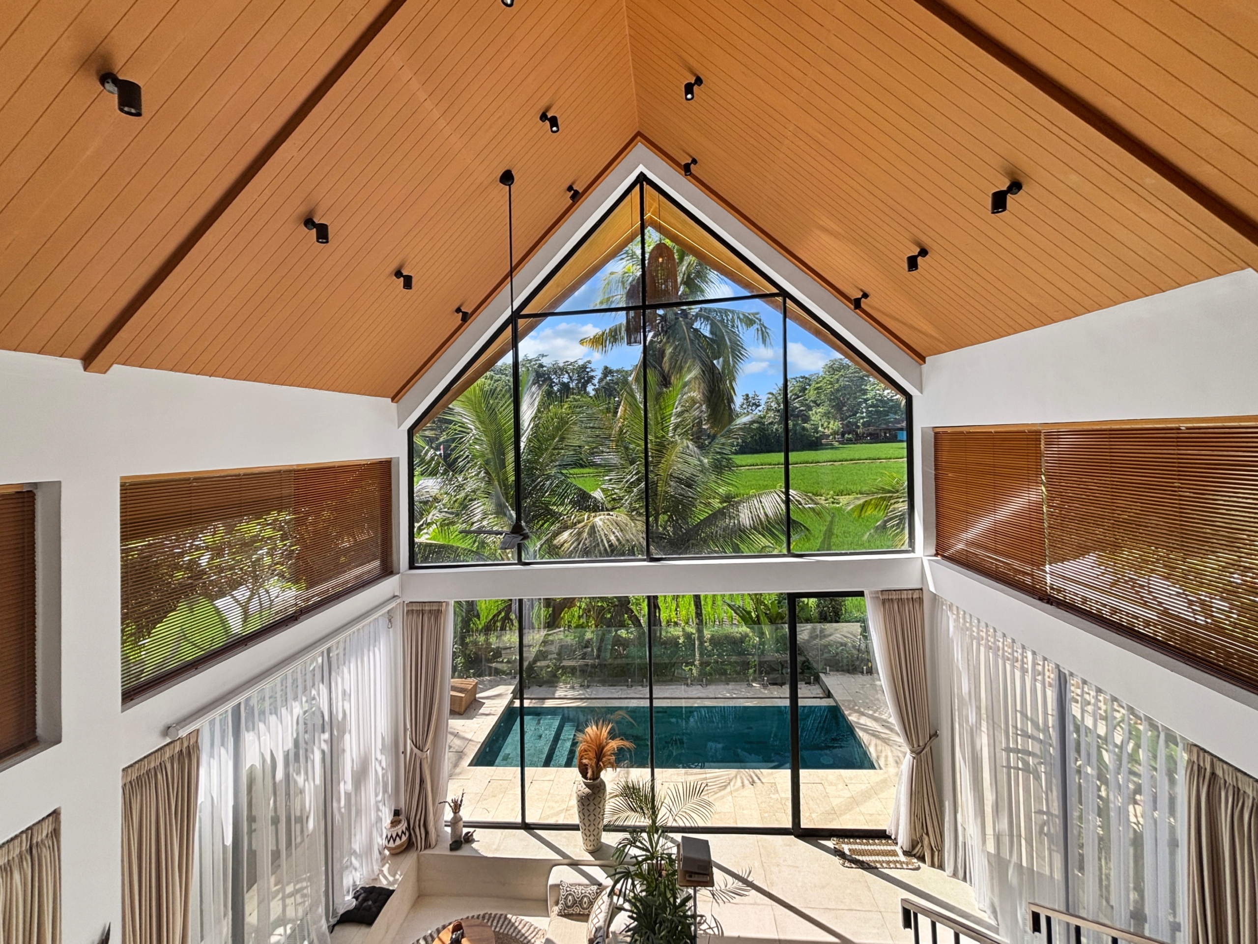 1201478c4c8224a3f95f89027305a56563e979fb Interior view of a double-height living area featuring a wooden vaulted ceiling, black-framed triangular window, and palm trees outside