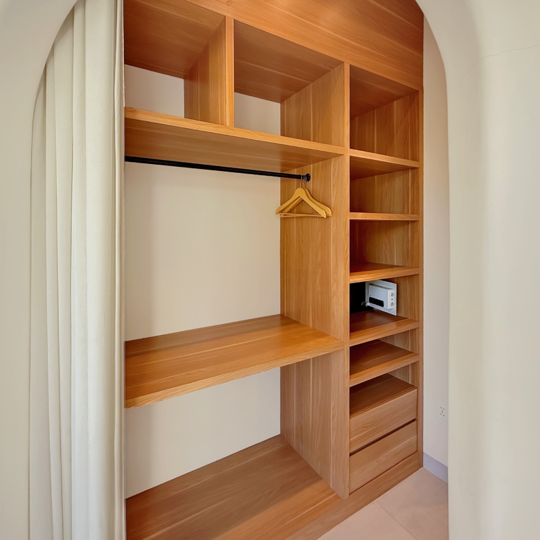 Open wooden wardrobe with multiple shelves and a metal hanging rod, a wooden clothes hanger resting on the rod, cream walls and curtain beside.