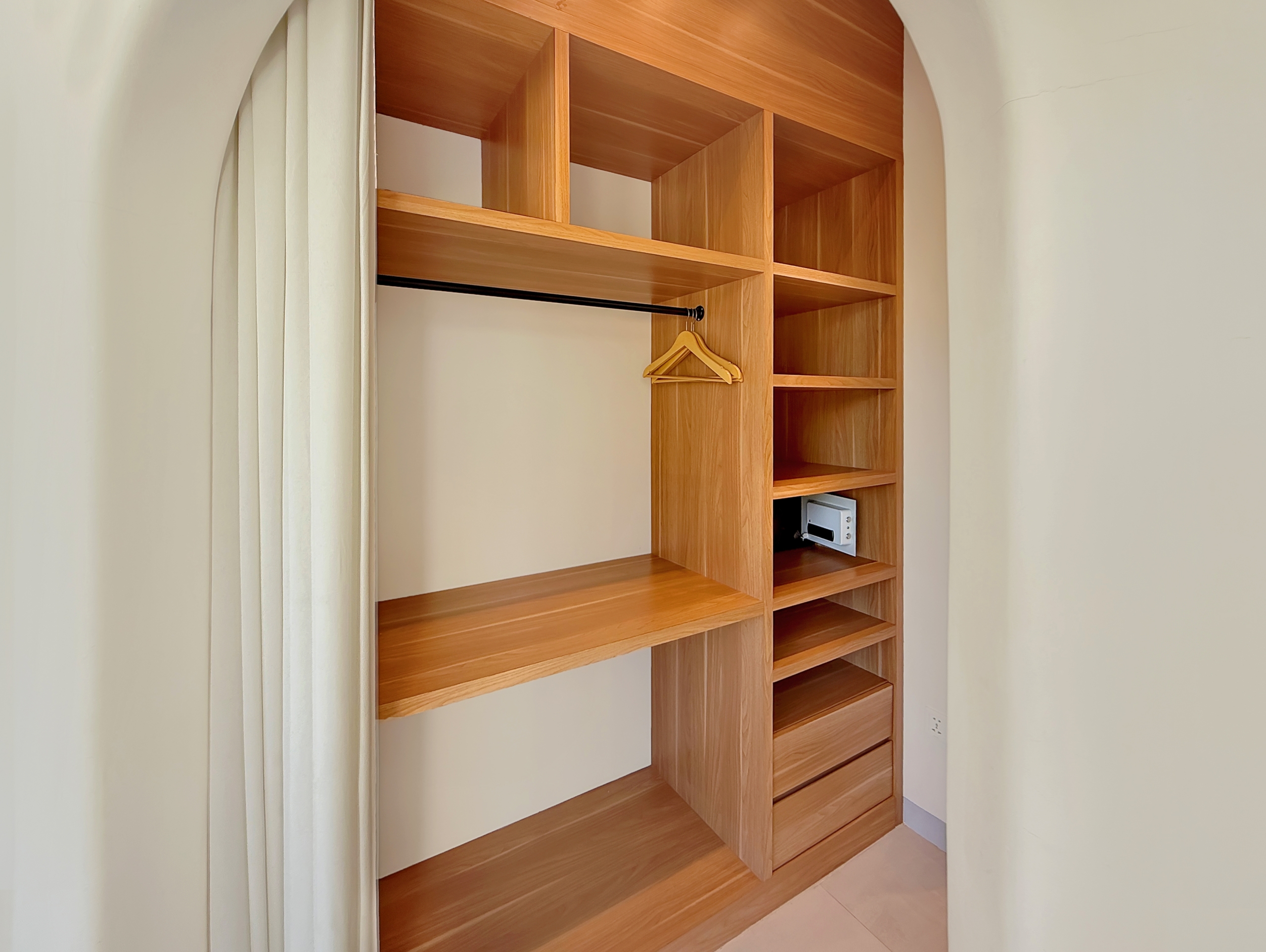 13af71b41a78cd8baa9d2e6ed164483fc6828776 Open wooden wardrobe with multiple shelves and a metal hanging rod, a wooden clothes hanger resting on the rod, cream walls and curtain beside.