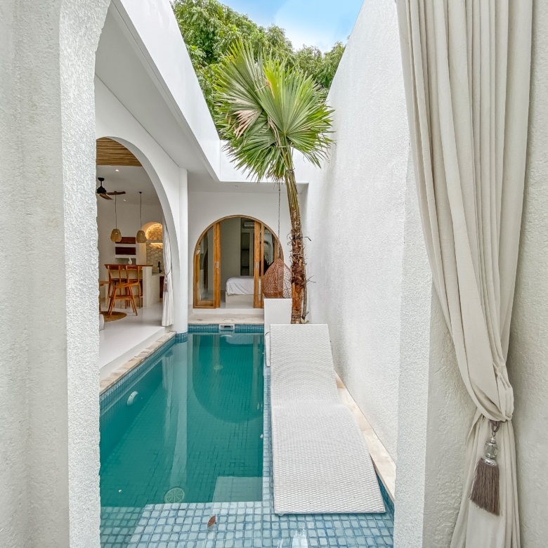 White stucco courtyard with a narrow blue-tiled pool, palm tree, and arched doorways leading inside, sunlit and airy