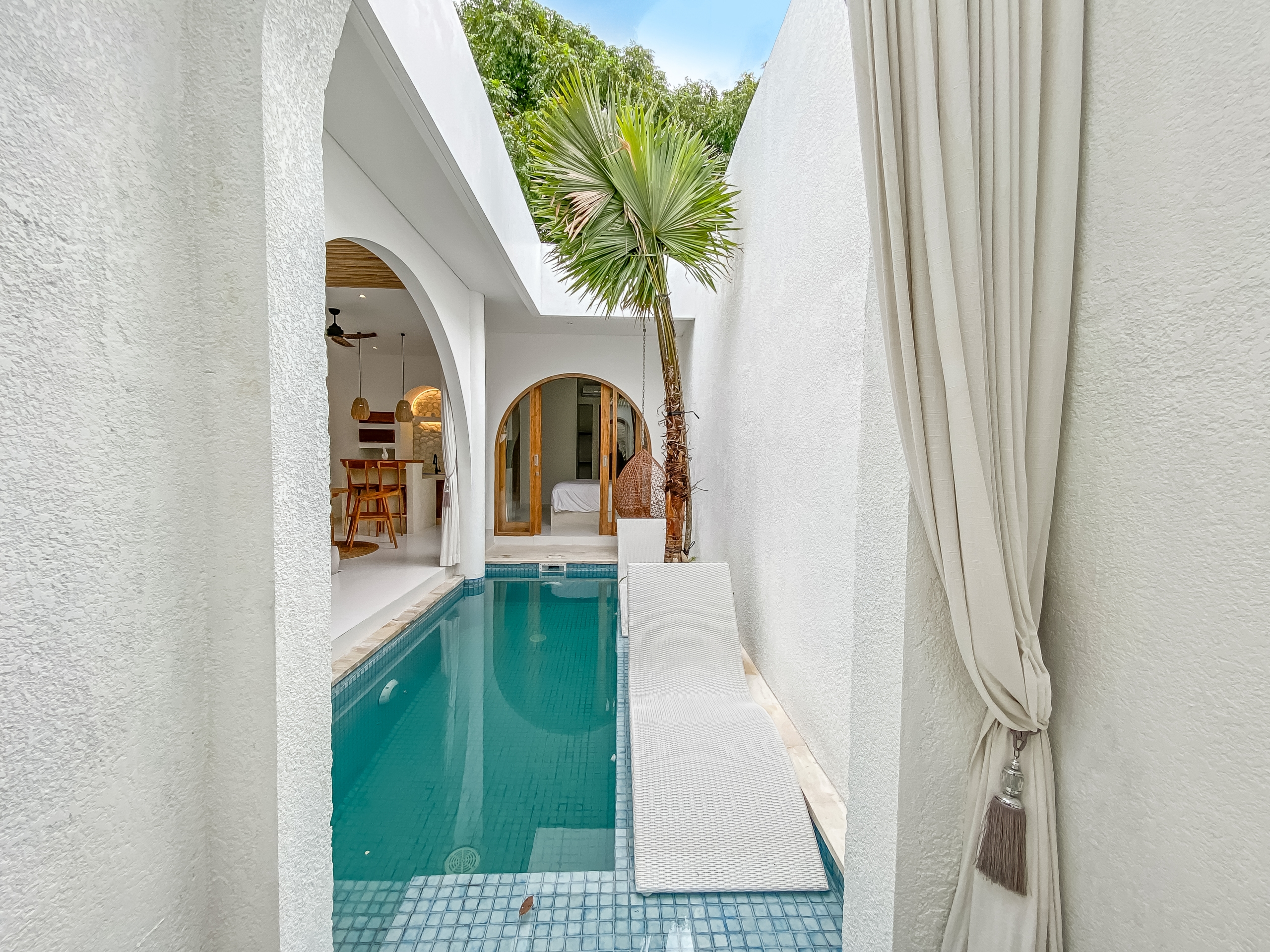1442a5a1d64b32001d92d76bade550ef45b03dfb White stucco courtyard with a narrow blue-tiled pool, palm tree, and arched doorways leading inside, sunlit and airy