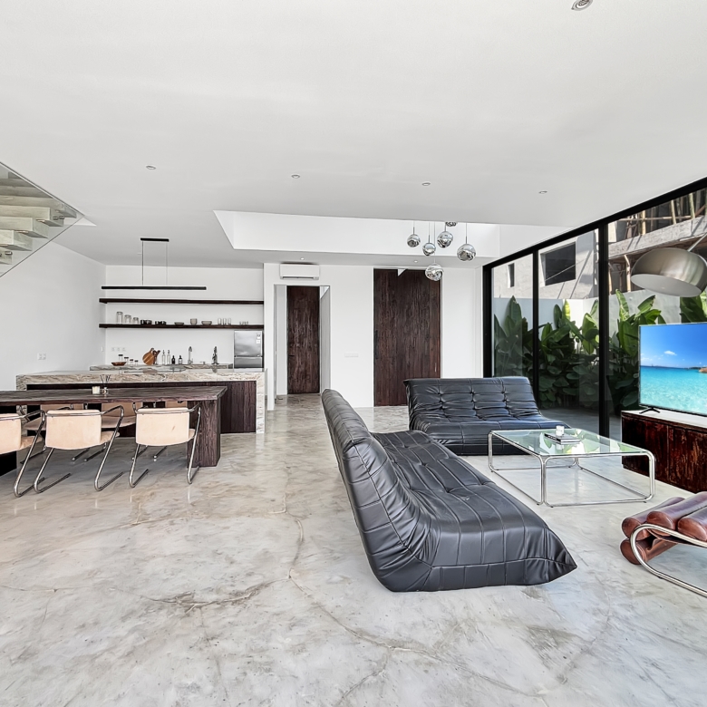 Open‑concept living/dining area with a glass‑walled staircase, marble floor, dark wood dining table, leather lounge chairs, and a large TV showing a tropical beach scene.