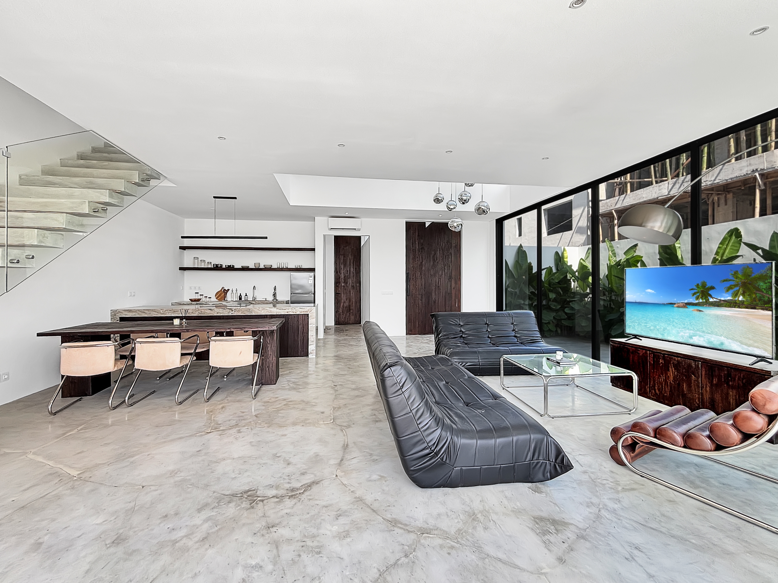 18089ad97993e77bcd2c99a28c8e38b8d84c2824_6781-6a1d7baddc487 Open‑concept living/dining area with a glass‑walled staircase, marble floor, dark wood dining table, leather lounge chairs, and a large TV showing a tropical beach scene.