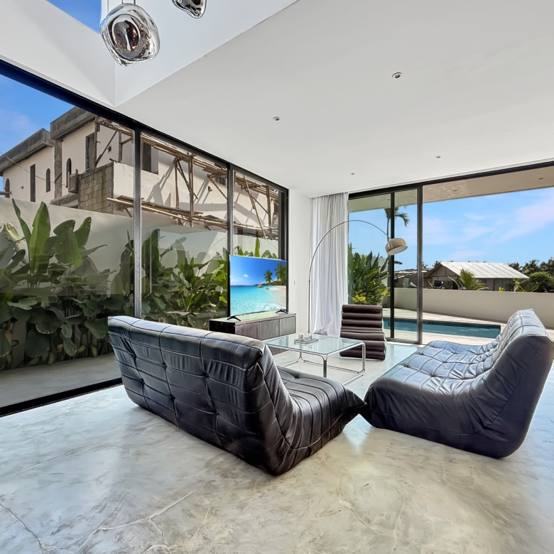 Open living room with black leather lounge chairs around a glass coffee table, large glass walls, and a backyard pool with blue sky.