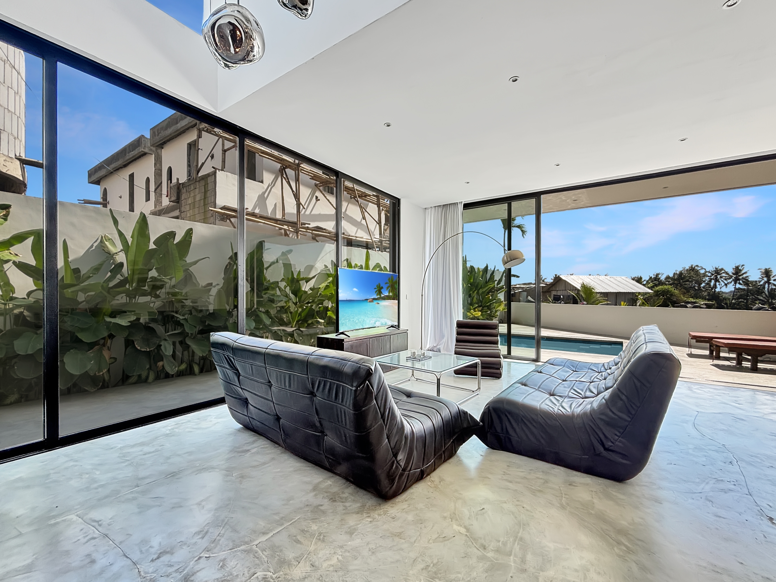 1e49646cabf448ff362fa81b7349490af4eb582b_6813-6a1d7bad896ab Open living room with black leather lounge chairs around a glass coffee table, large glass walls, and a backyard pool with blue sky.