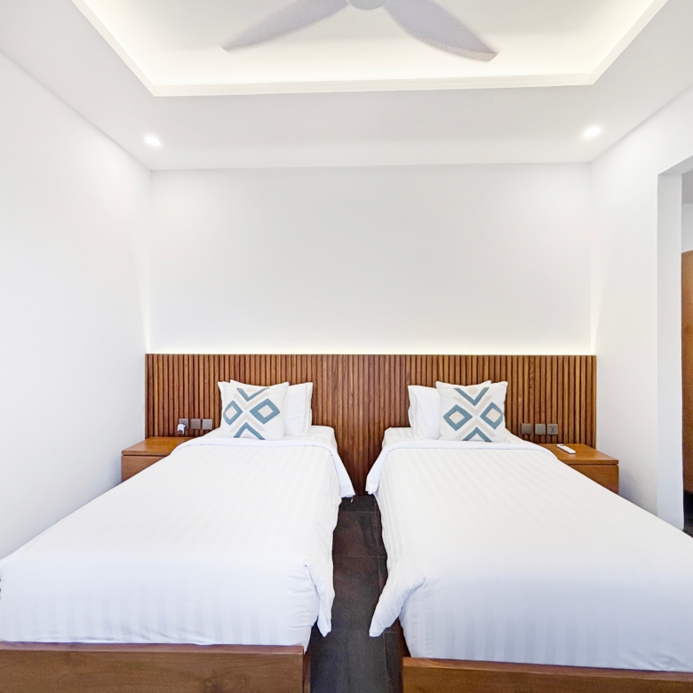 Two twin beds with white linens and a wooden slatted headboard, each with blue geometric pillows in a bright hotel room.