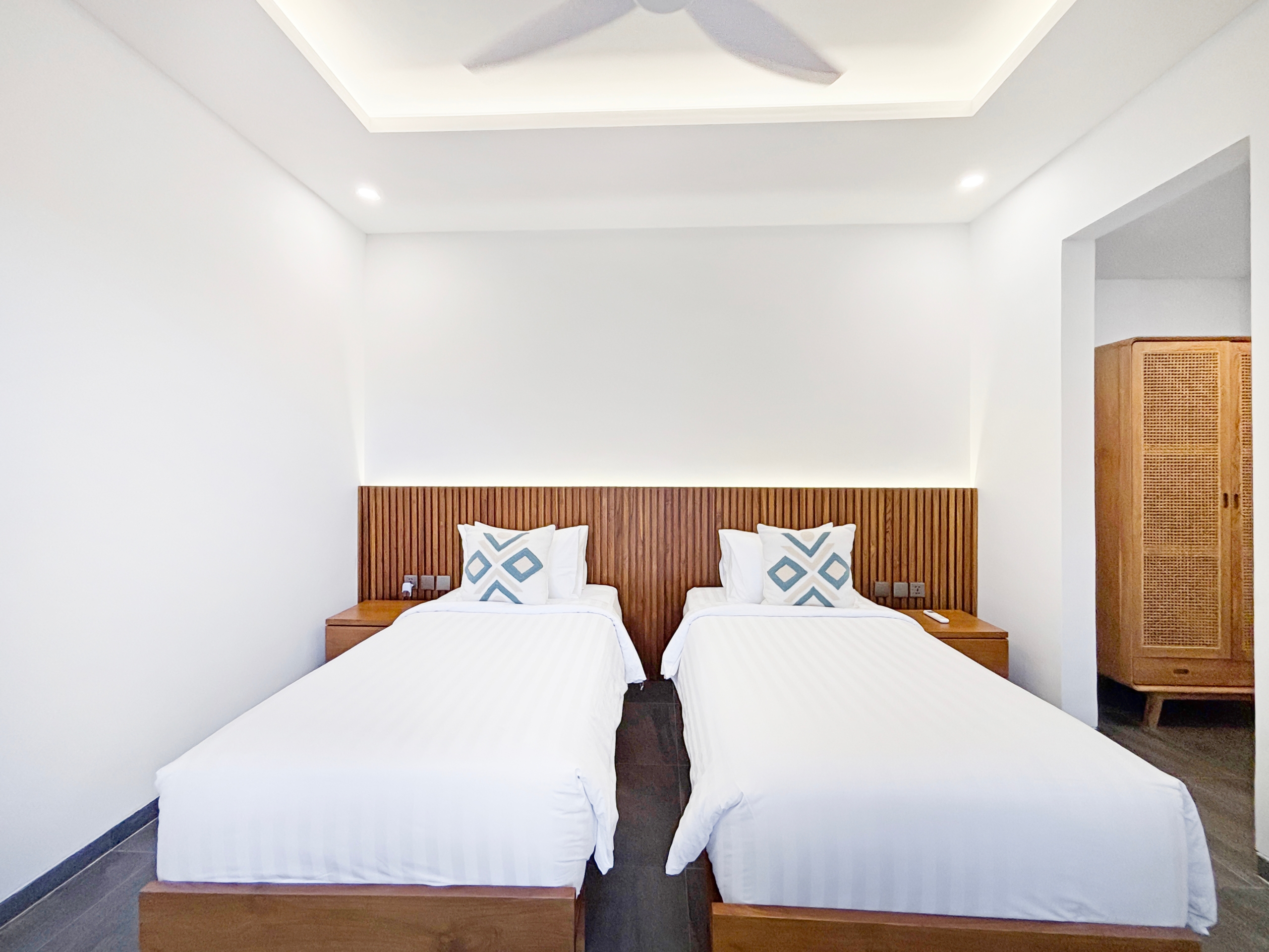 1edf04f557c4e8d76eea4b052a311b2b2ac6da26 Two twin beds with white linens and a wooden slatted headboard, each with blue geometric pillows in a bright hotel room.