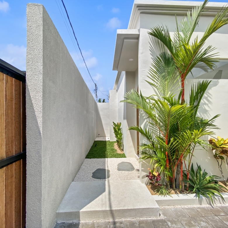Modern white building with a slim walkway between walls, lush tropical plants on the right, and a wooden fence on the left under a clear blue sky.