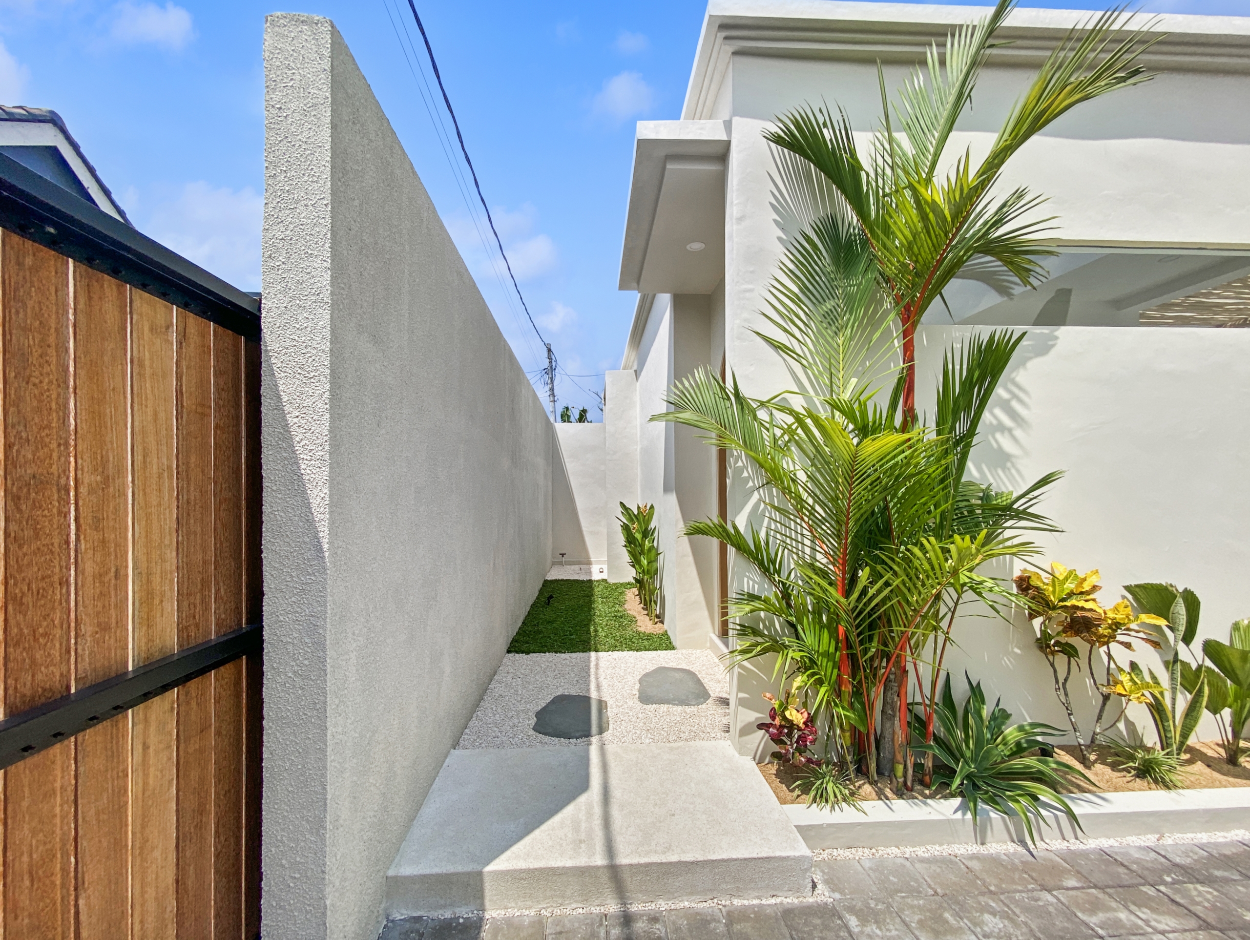 1fe0dd63dcffb6506a59c28c94e99f1415e985eb_2285-68d5cfae2b8e6 Modern white building with a slim walkway between walls, lush tropical plants on the right, and a wooden fence on the left under a clear blue sky.