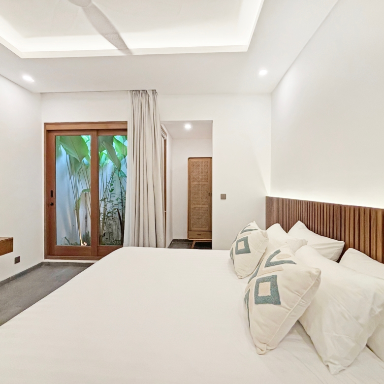 Bright hotel bedroom with white walls, a wooden slatted headboard, white bedding, and decorative pillows; glass door opens to lush tropical plants outside.