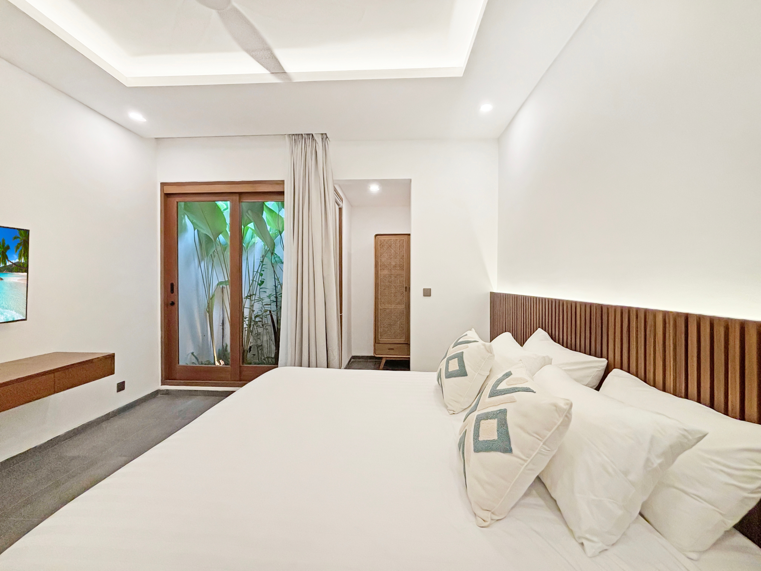 22accad64d9432bdf04db220e7d3bb671be6527b Bright hotel bedroom with white walls, a wooden slatted headboard, white bedding, and decorative pillows; glass door opens to lush tropical plants outside.