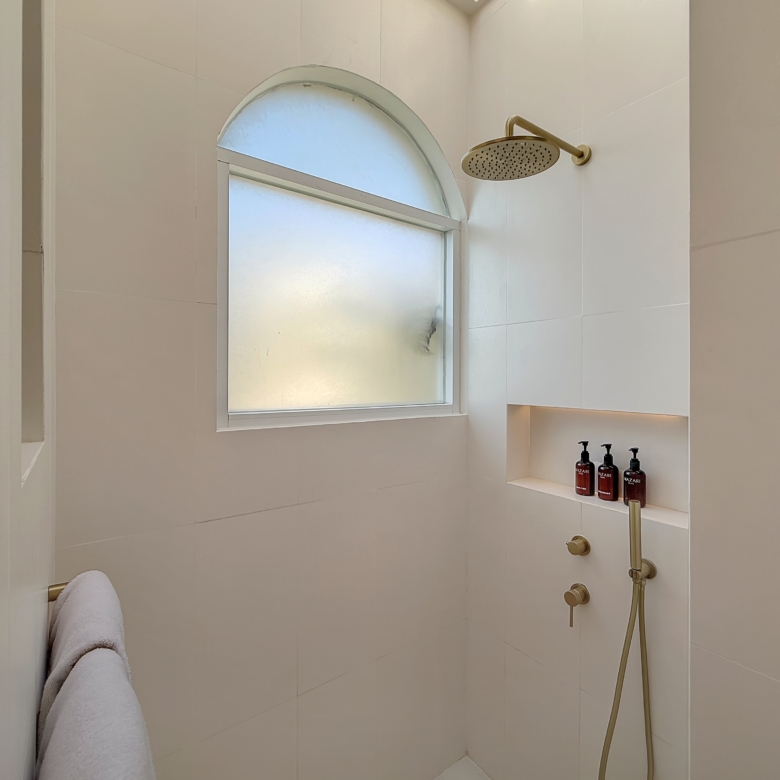 White-tiled shower stalls with a round rainfall showerhead and a curved arched window, plus a built-in shelf holding brown bottles of bath products.
