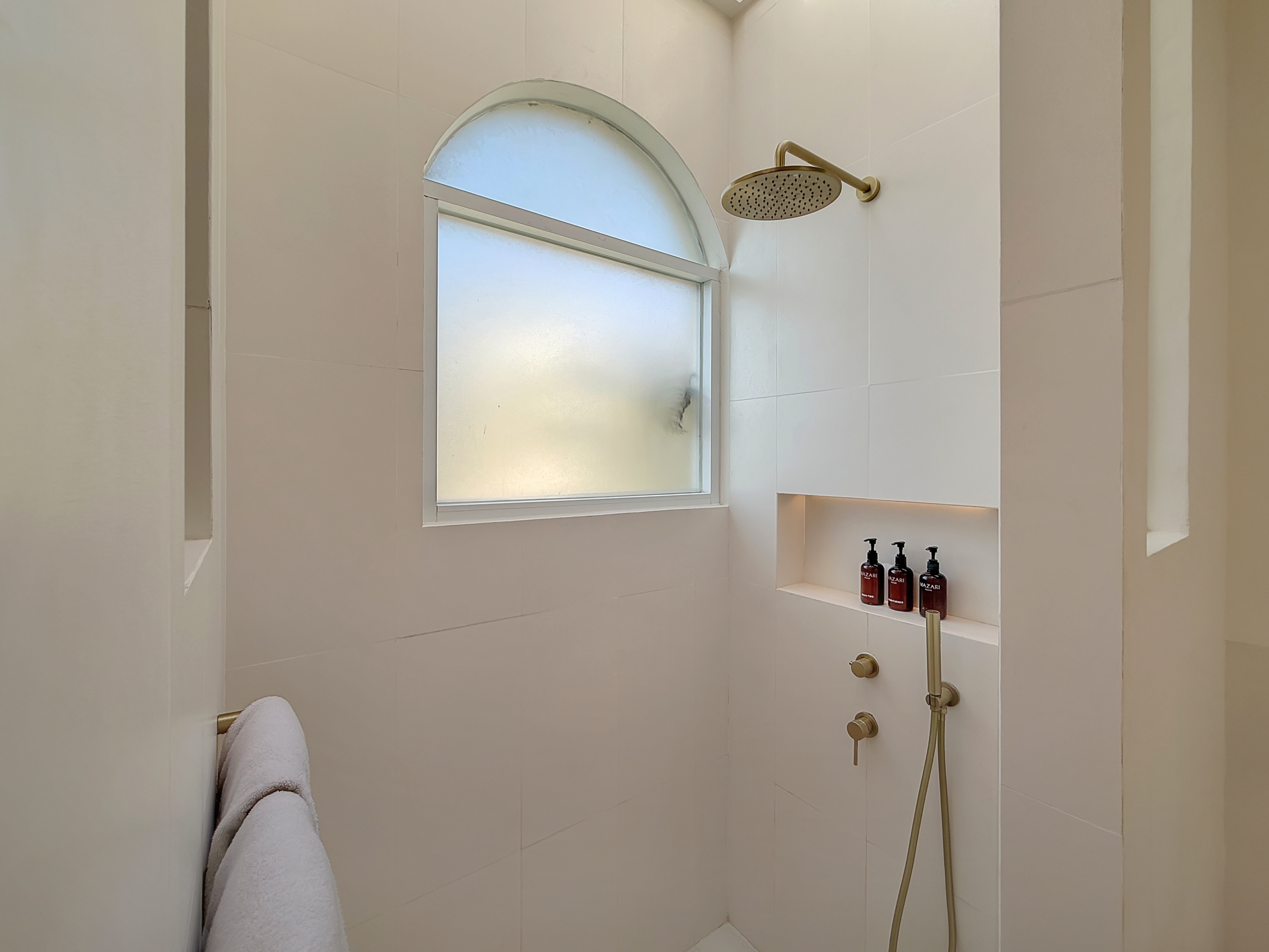 22ef1ee9ba8d5f70c06be79de341e1bab59e3a2b White-tiled shower stalls with a round rainfall showerhead and a curved arched window, plus a built-in shelf holding brown bottles of bath products.