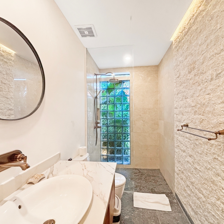 Bathroom with a double sink vanity, round mirror, bronze faucet, and beige marble countertop; glass block window at the far end adds privacy and light.