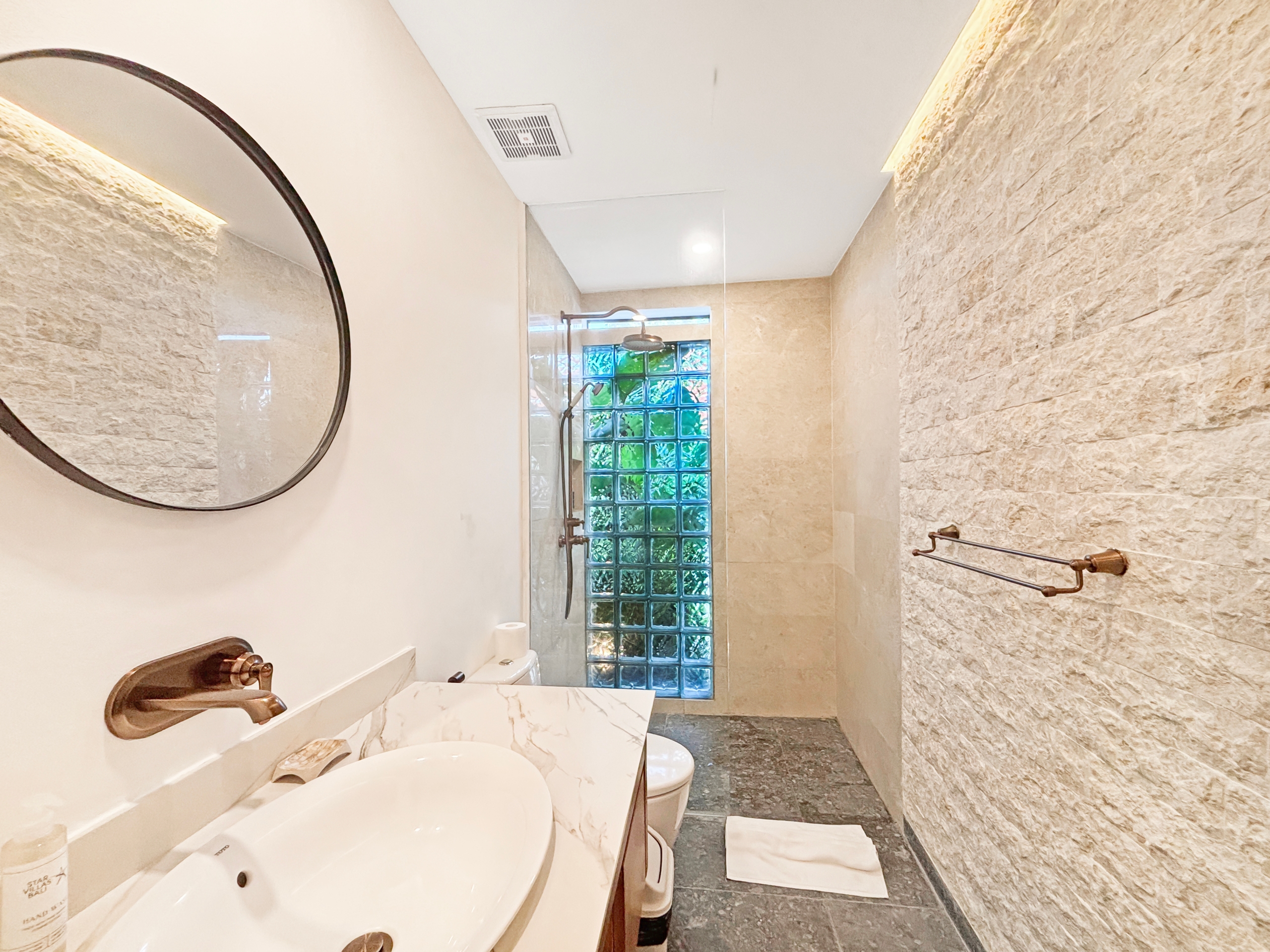 2a6f6440982dc8f8e6c4f3e7f2c22bd9a55850f0 Bathroom with a double sink vanity, round mirror, bronze faucet, and beige marble countertop; glass block window at the far end adds privacy and light.
