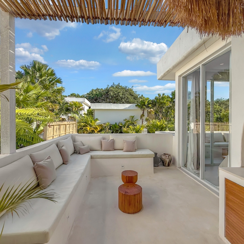Outdoor lounge area with a white sectional sofa and beige cushions, two wooden stump tables, glass sliding doors, and tropical plants under a thatched roof in bright sunshine.