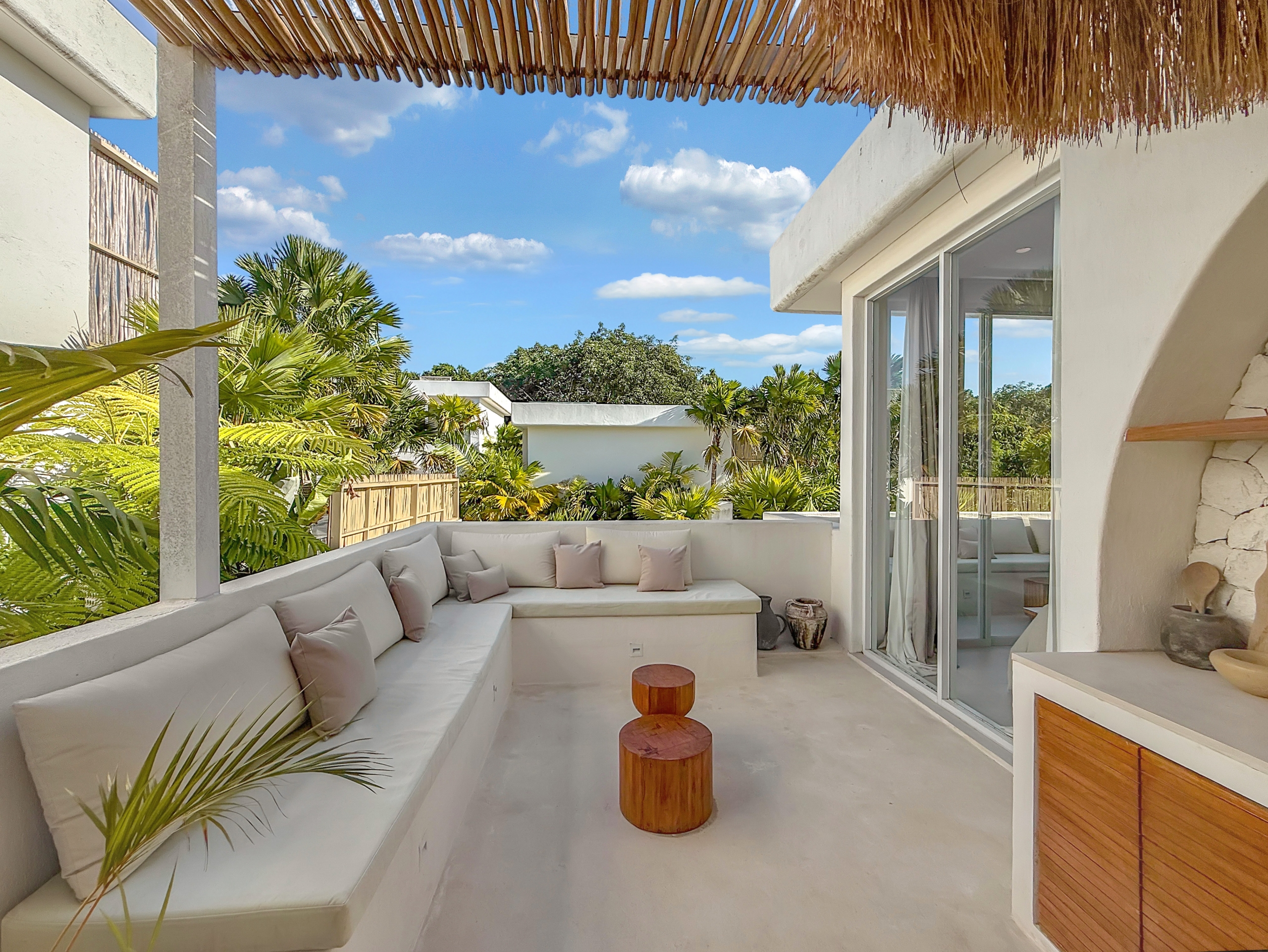 2ab3c7373310e081e8935a978a1a75a99349691d Outdoor lounge area with a white sectional sofa and beige cushions, two wooden stump tables, glass sliding doors, and tropical plants under a thatched roof in bright sunshine.