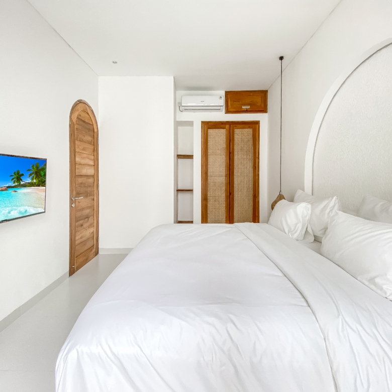 Bright bedroom with two white-titted beds (duvet-covered) and a wall-mounted TV showing a tropical beach scene, wooden arched door nearby, and white walls.
