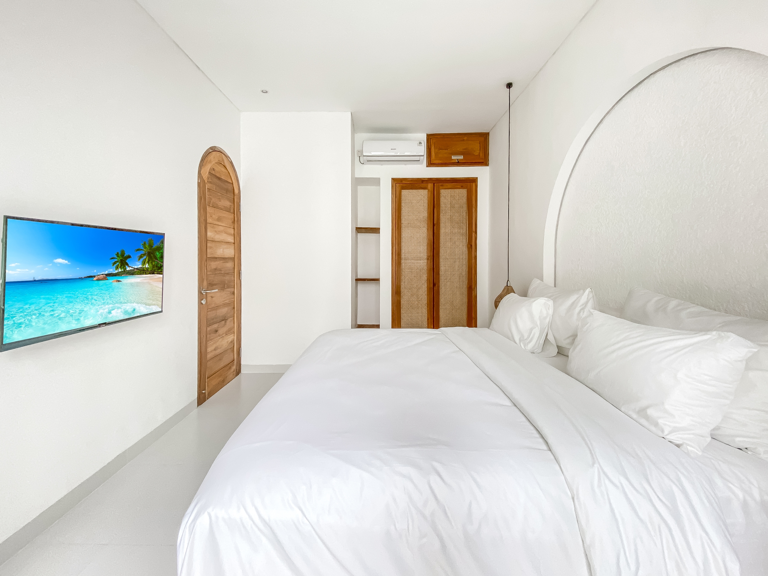 2b96470a999acabe463065f0ce4f91720f51ad18 Bright bedroom with two white-titted beds (duvet-covered) and a wall-mounted TV showing a tropical beach scene, wooden arched door nearby, and white walls.