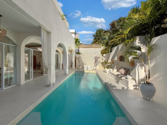 White modern villa courtyard with a rectangular turquoise pool, arches, glass doors, and draped curtains along a sunny patio.